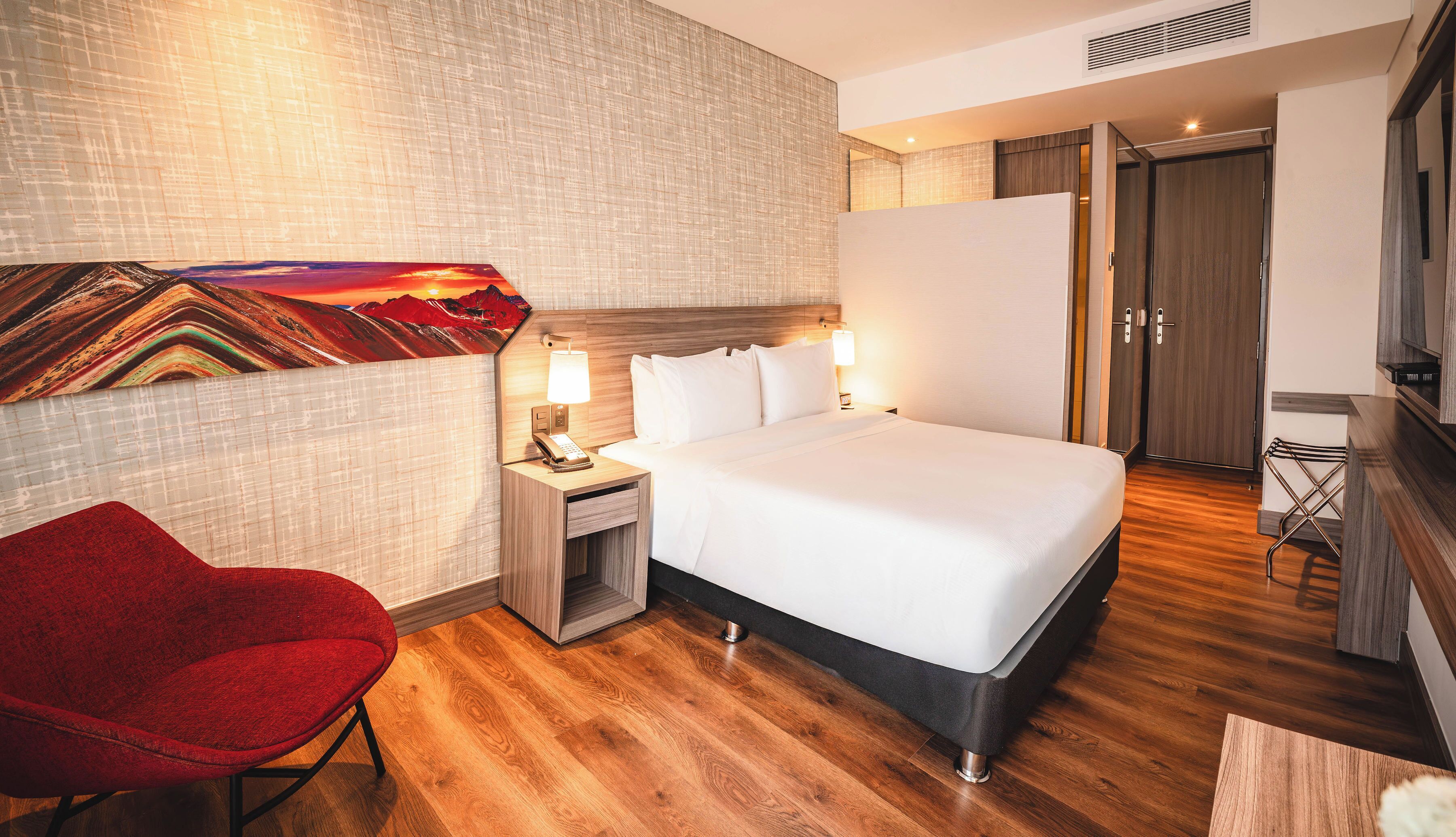 A hotel room with a bed, a red chair, a desk with a lamp, and a painting on the wall.
