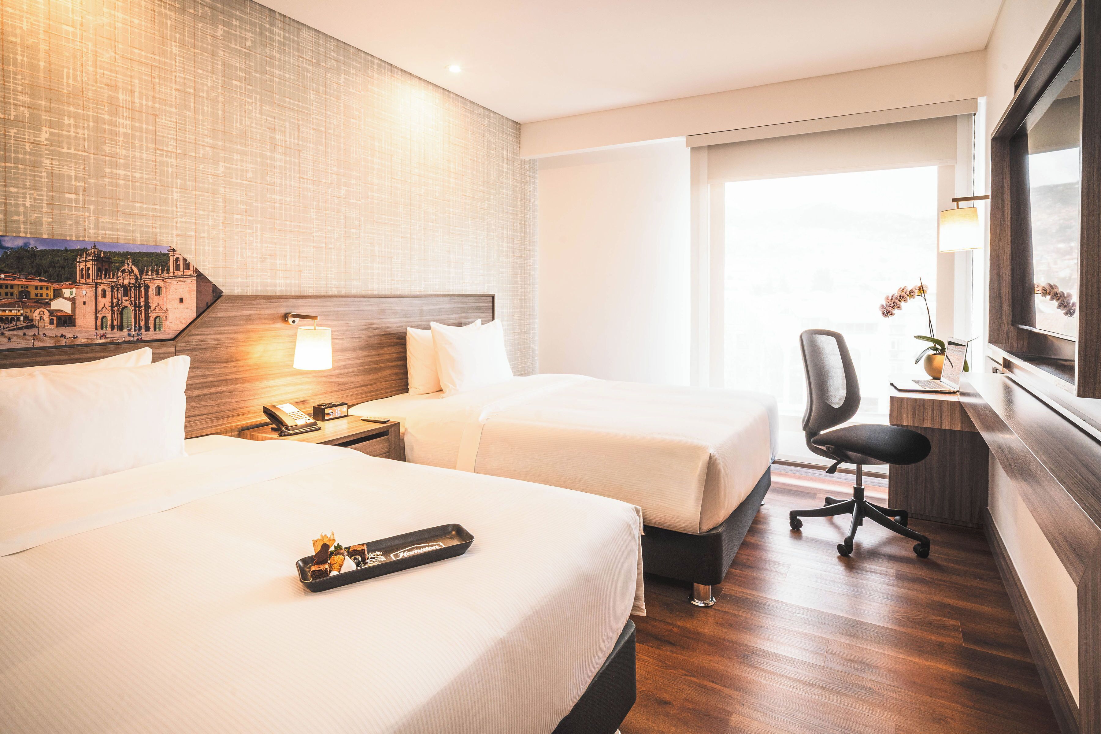 A hotel room with two beds, a desk, a chair, a nightstand, and a large window.