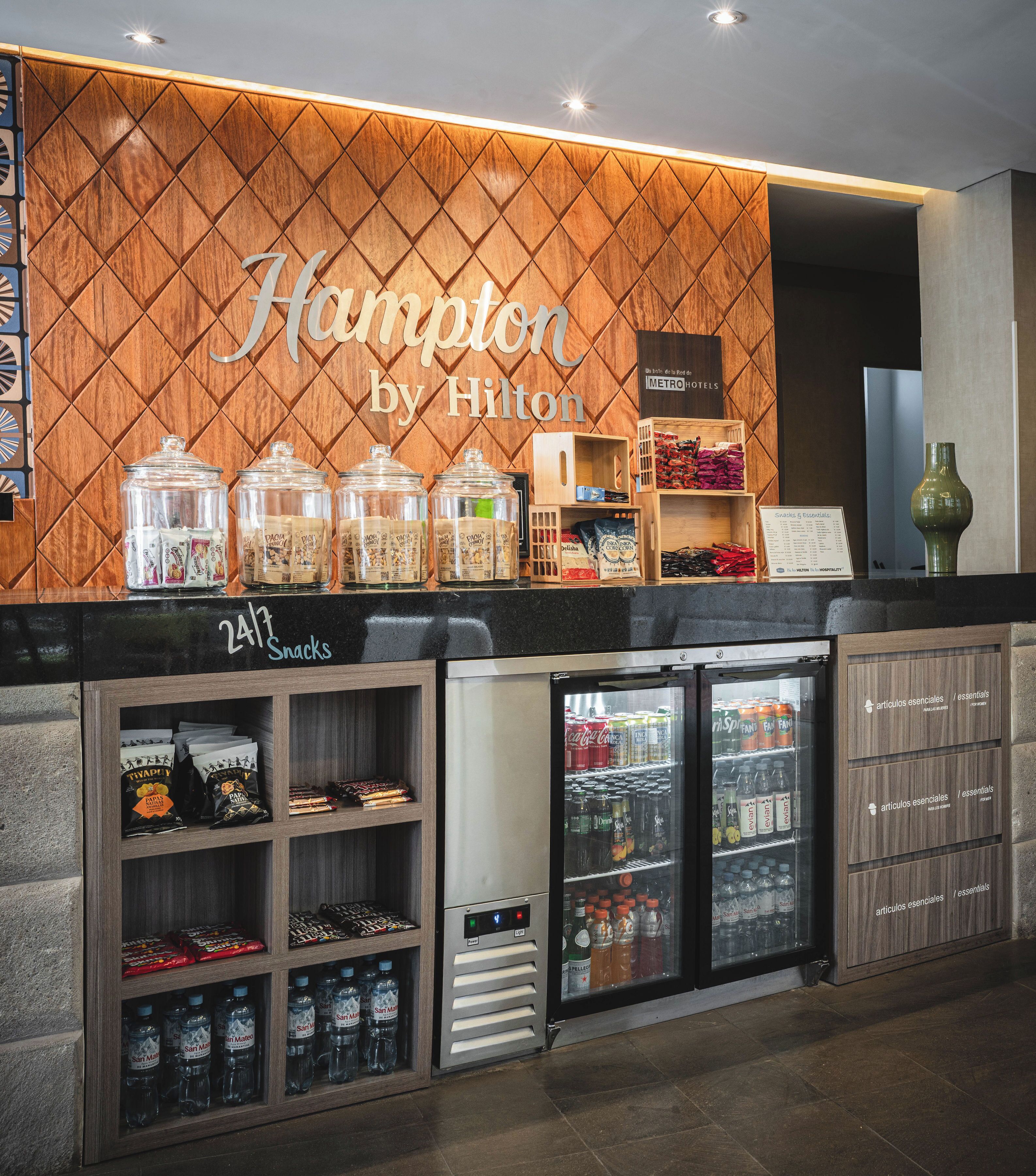 A hotel reception area with a wooden wall displaying the name 'Hampton by Hilton'. There are glass jars with snacks, a refrigerator stocked with beverages, and a sign for '24/7 Snacks'.