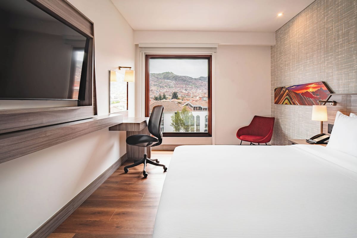 A hotel room with a large bed, a desk with a chair, a TV, a red chair, and a view of a cityscape through a window.