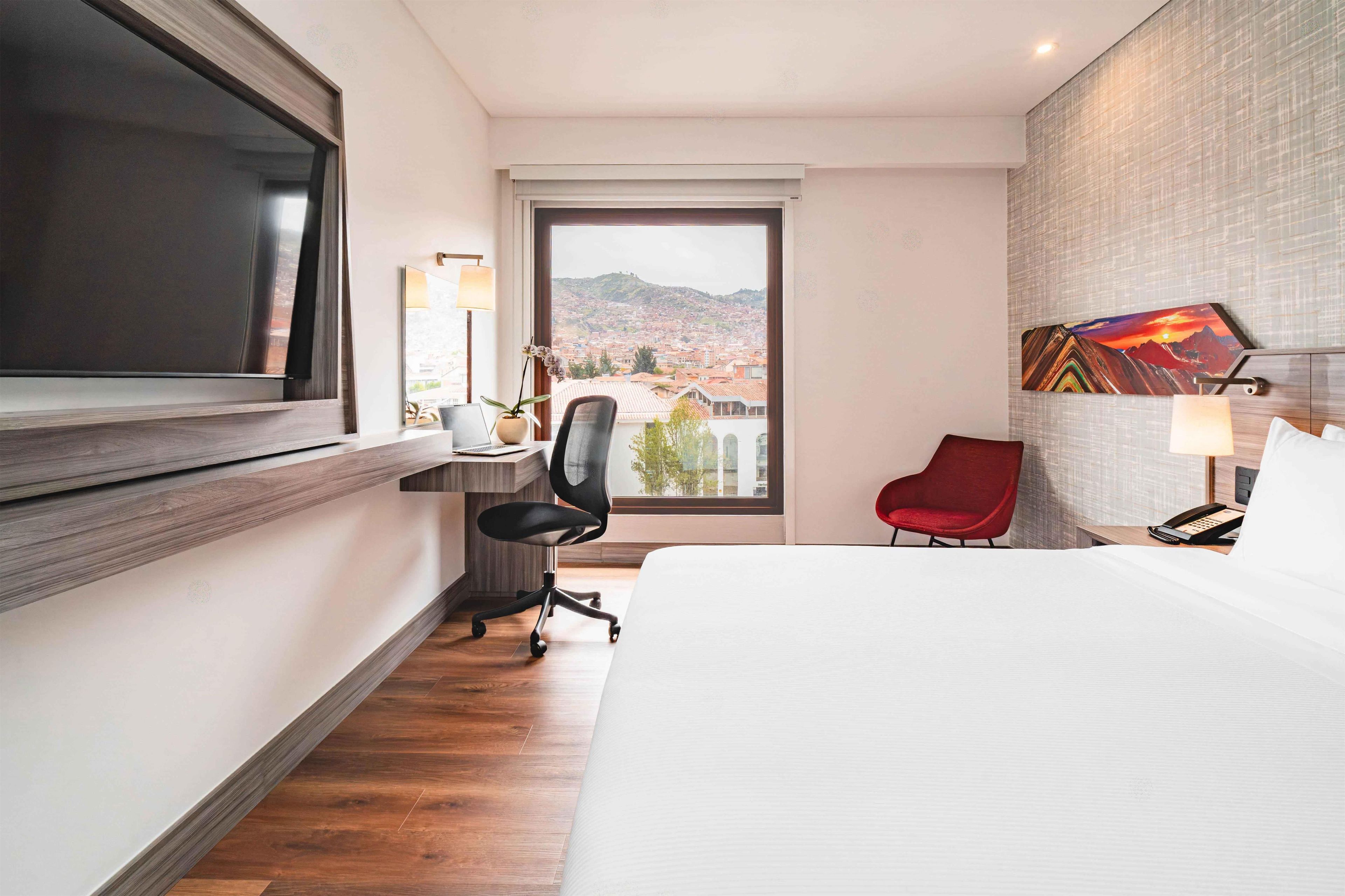 Habitación de hotel con cama, escritorio, silla, televisor, ventana con vista a la ciudad y un cuadro en la pared.