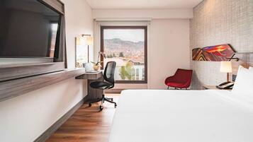 Habitación de hotel con cama, escritorio, silla, televisor, ventana con vista a la ciudad y un cuadro en la pared.