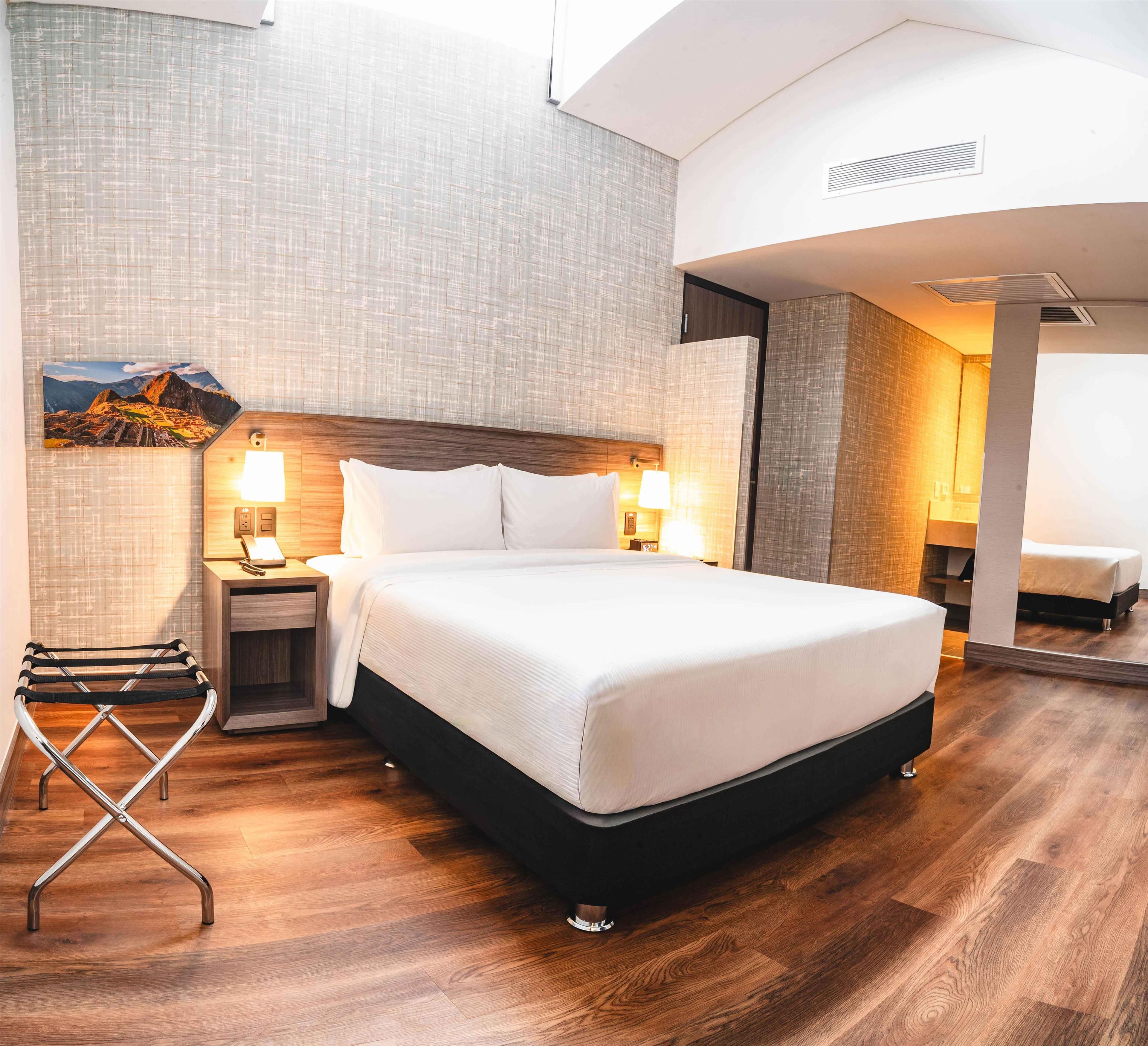 Een hotelkamer met een groot bed, houten vloer, nachtkastje met lamp, een aan de muur hangend schilderij en een spiegel.