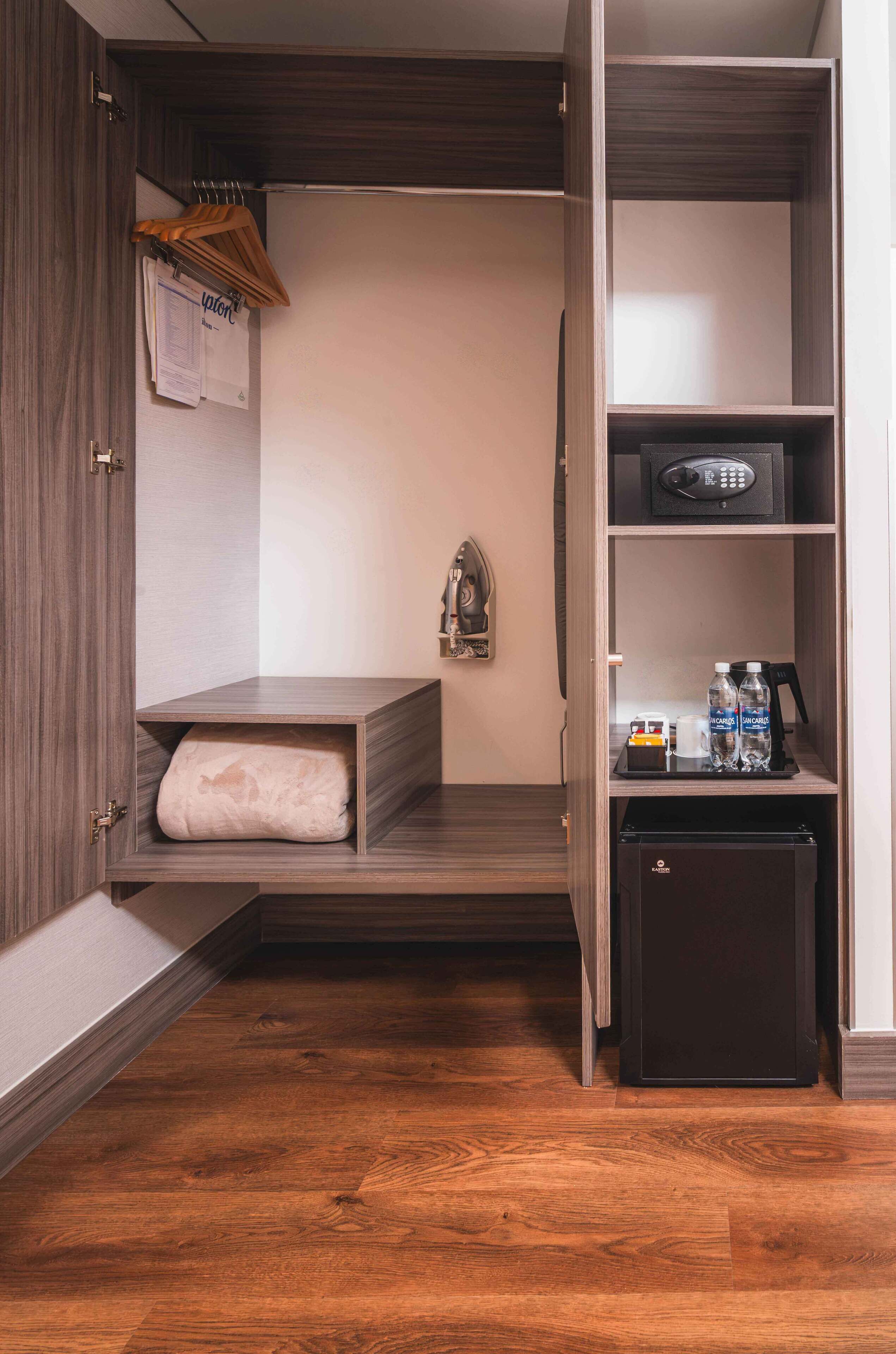 A compact hotel room with a built-in wardrobe, a small seating area, a minibar, and a desk with a telephone and bottled water.
