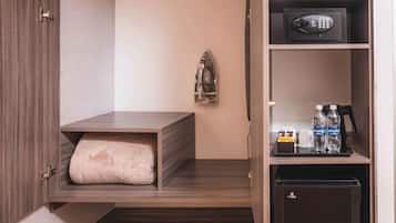A compact hotel room with a built-in wardrobe, a small seating area, a minibar, and a desk with a telephone and bottled water.