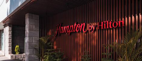 A hotel entrance with a red facade and the name "Hampton by Hilton" in large letters.