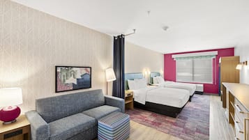 A hotel room with a grey sofa, a small table with a lamp, a bed with two pillows, a nightstand, a wooden dresser, and a large window with a curtain.