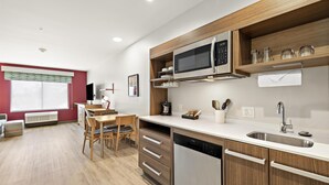 A modern kitchen with wooden cabinets, a stainless steel dishwasher, and a microwave. There is a dining area with a table and chairs, and a window with blinds. - Home2 Suites by Hilton Riverside Downtown Convention Center (Riverside)