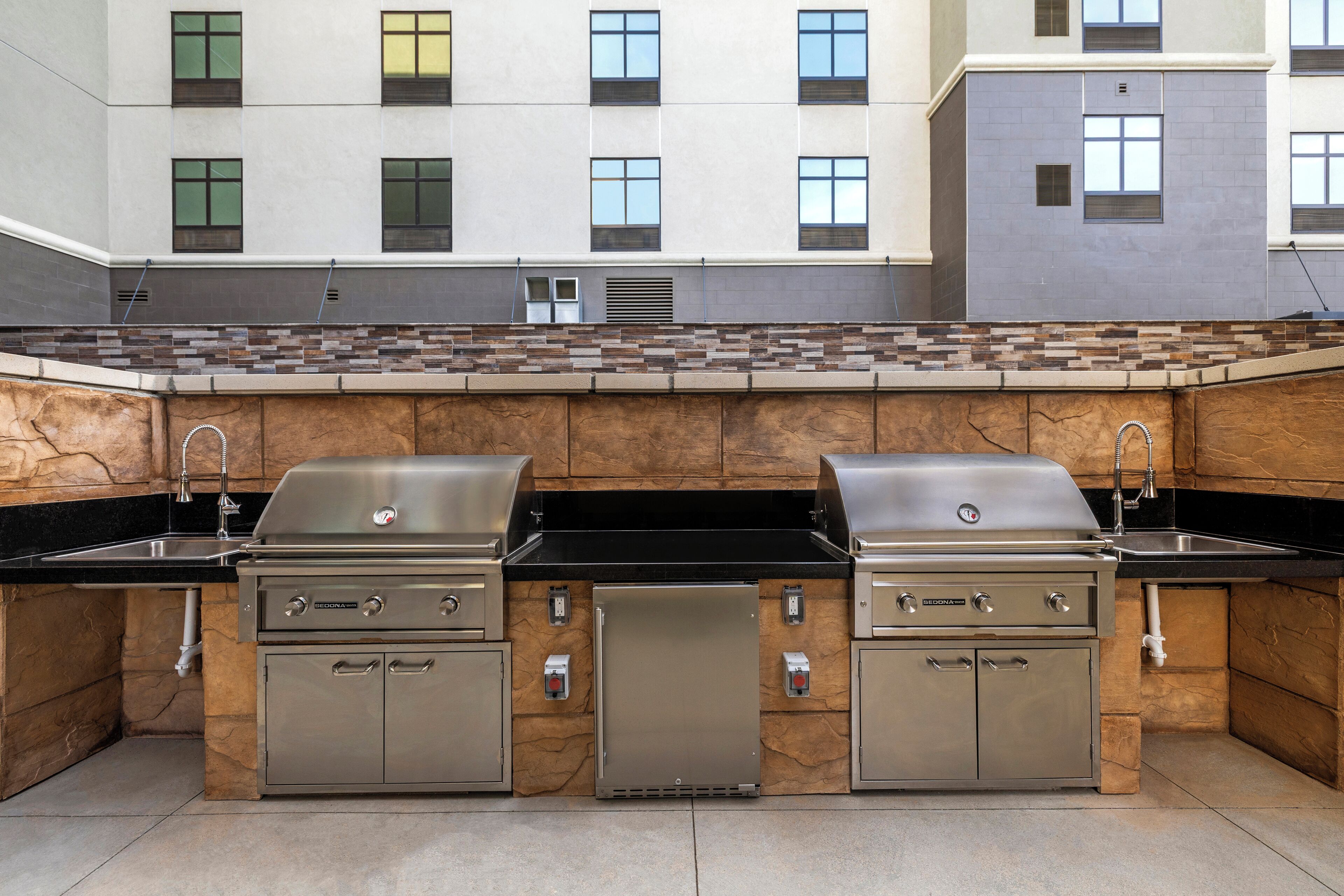 An outdoor kitchen area with two stainless steel grills, a sink, and a stone backsplash.