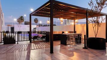 Un patio exterior con pérgola, mobiliario, zona de barbacoa y vistas a la ciudad.