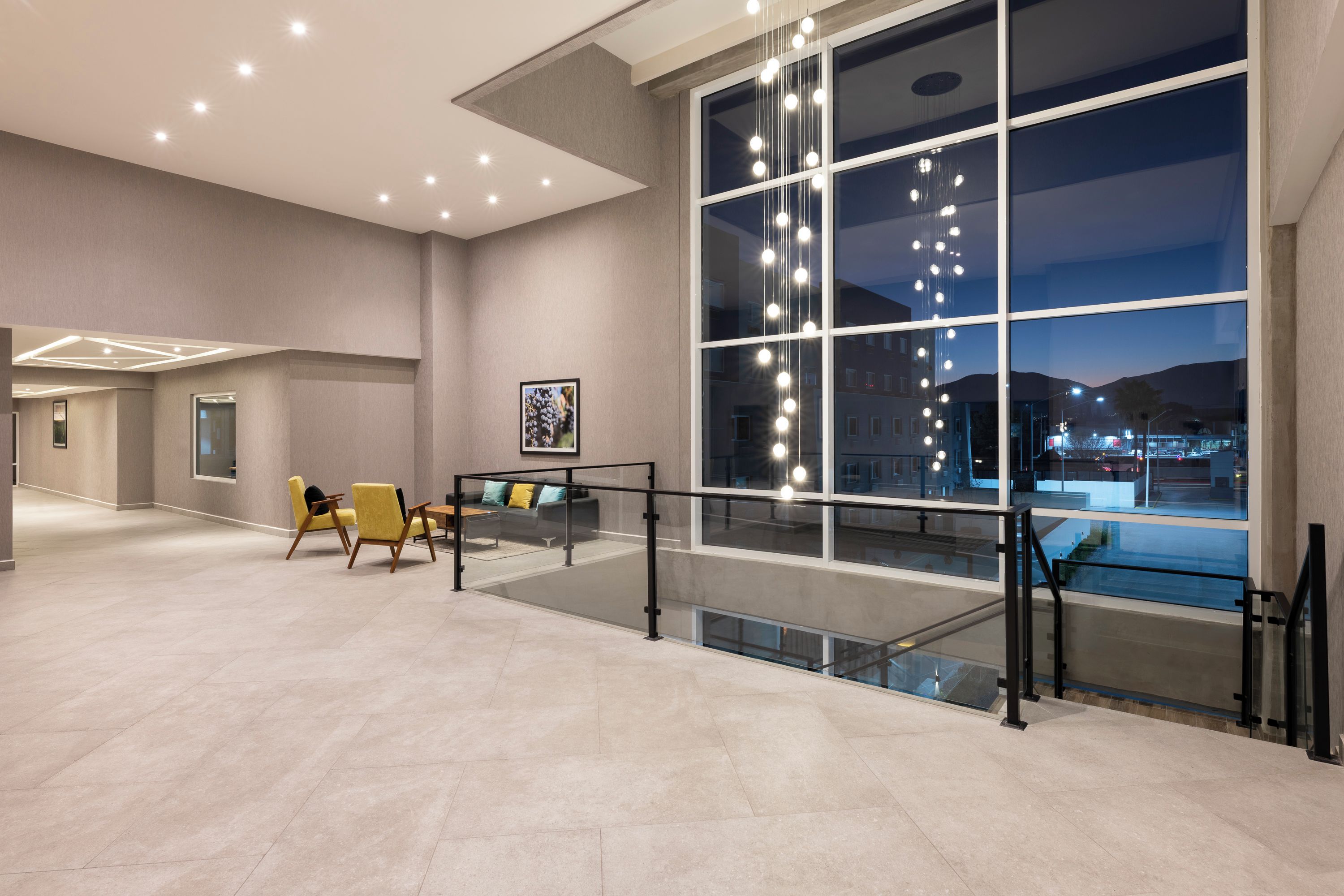 Een ruime lobby met een modern design, met een zithoek met gele stoelen, een glazen balustrade en een groot raam met uitzicht op de stad 's nachts.