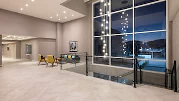Een ruime lobby met een modern design, met een zithoek met gele stoelen, een glazen balustrade en een groot raam met uitzicht op de stad 's nachts.
