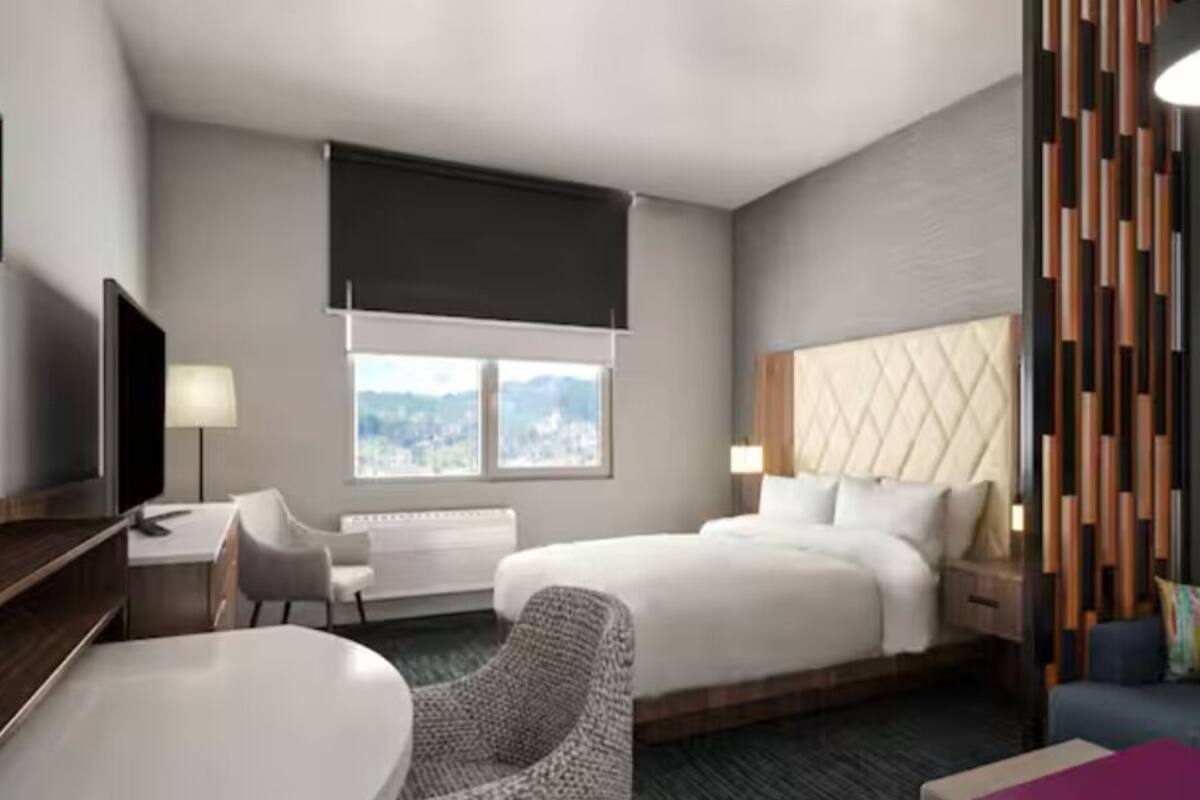 A modern hotel room with a large bed, a desk, a chair, and a sofa. There is a window with a view of the city.