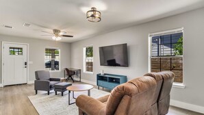 Un salon moderne avec un canapé en cuir marron, un fauteuil gris, une petite table ronde et un meuble bleu. Un téléviseur à écran plat est fixé au mur, et il y a des fenêtres équipées de stores.