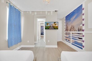 A hotel room with a bed, a TV, a window with a view of palm trees, and a wall with a snowy landscape.
