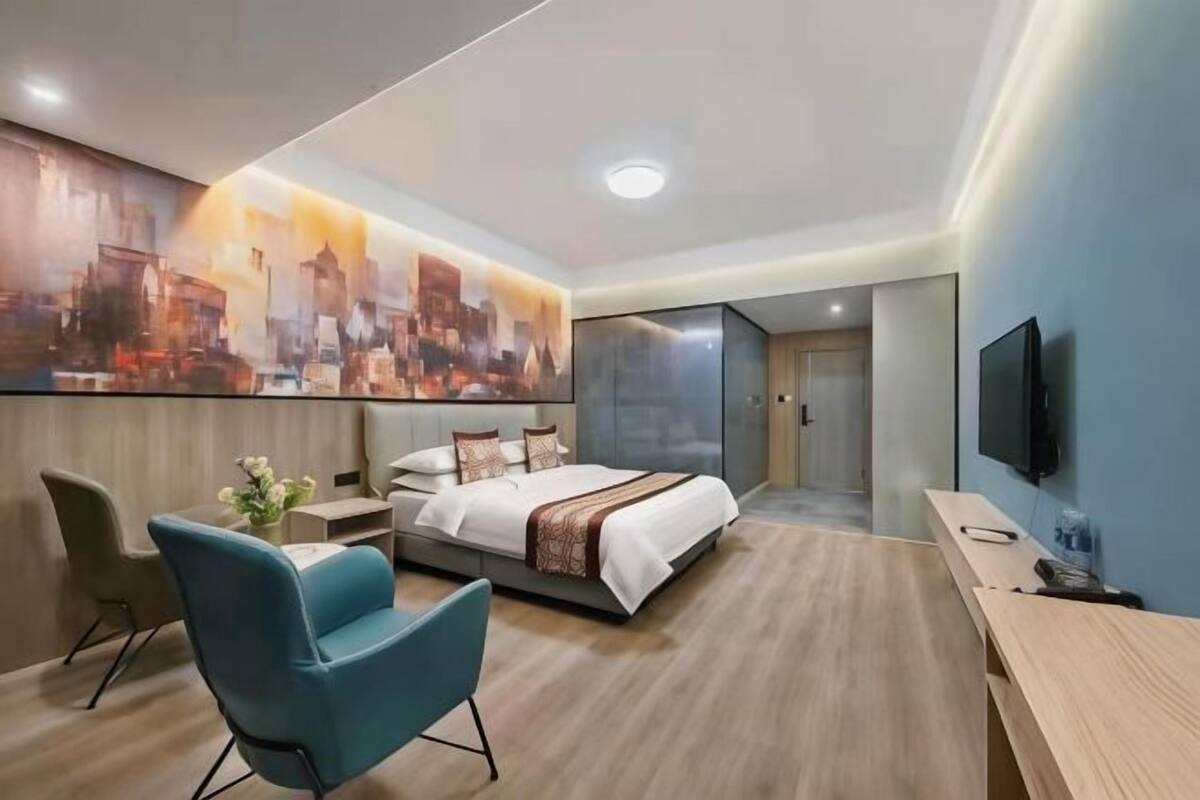 A modern hotel room with a large bed, a desk, a chair, a TV, and a cityscape painting on the wall.