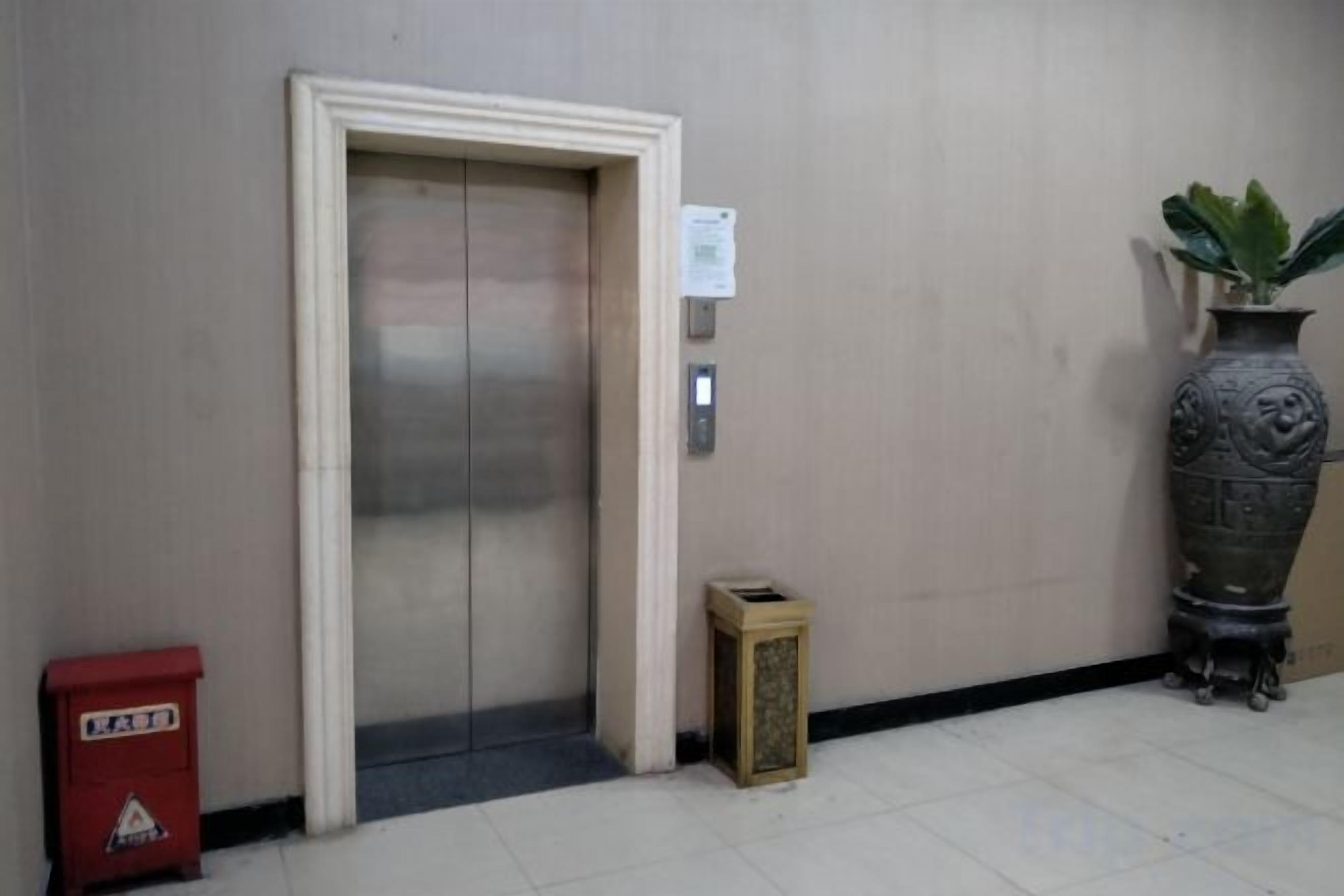 An elevator with a door, a small trash bin, a red trash bin, and a large decorative vase on the floor.