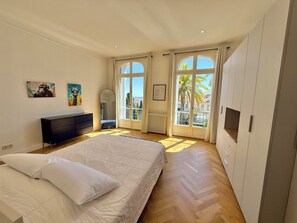 A bedroom with a bed, a dresser, a radiator, and a window with a view of palm trees. - Cannes Tassigny – Stylish - 2 Bedroom Apartment – Sleeps 5 – Free Parking (Cannes)