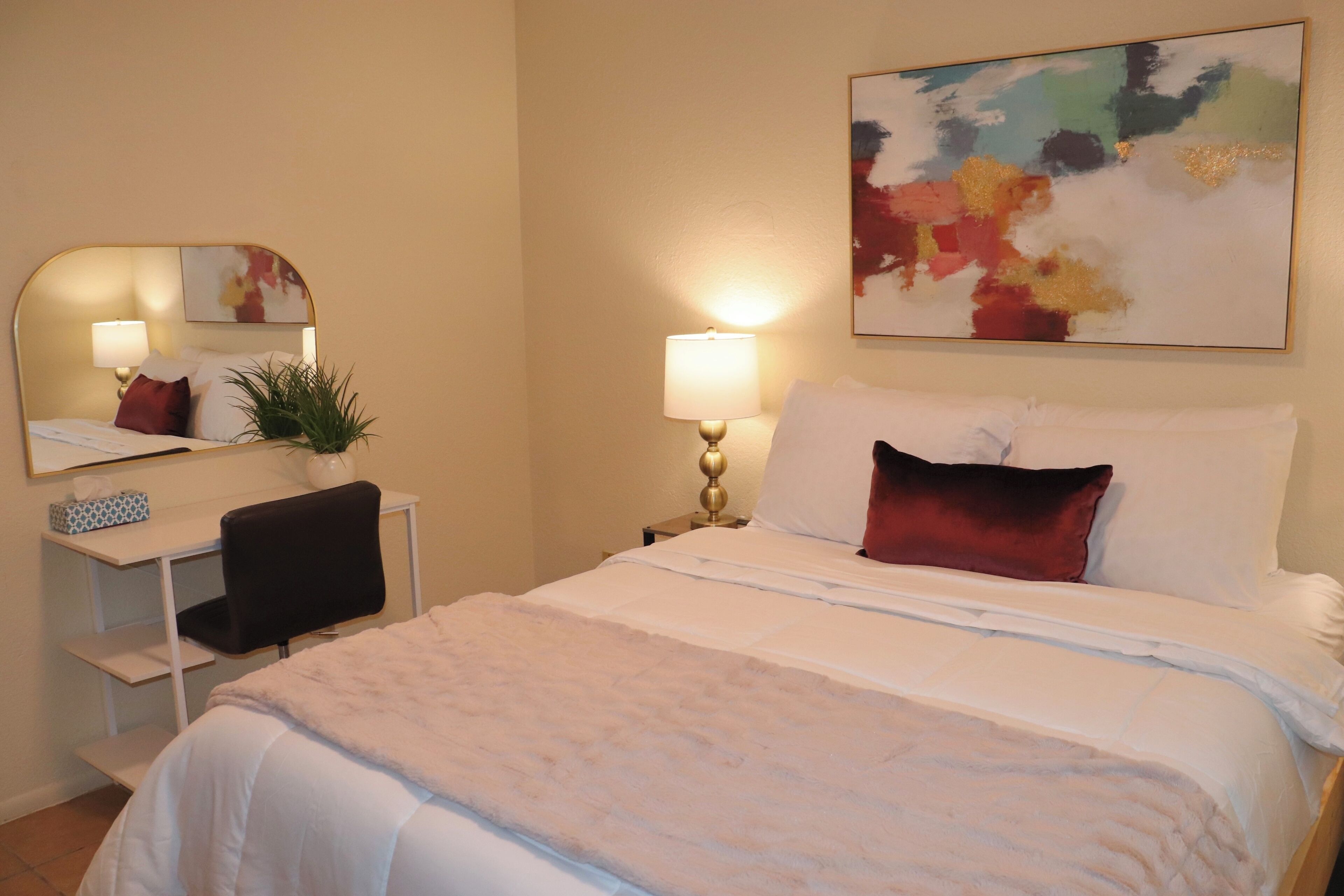 Habitación de hotel con cama, escritorio, silla, lámpara, espejo y un cuadro en la pared.