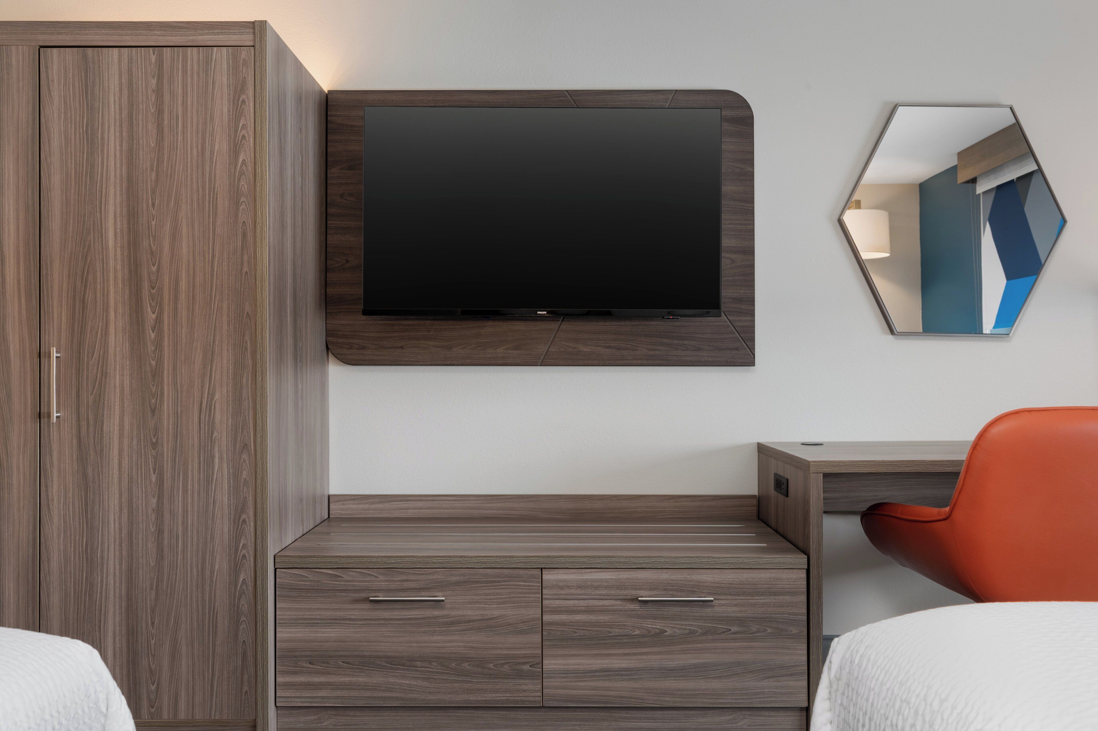 Moderni hotellihuone, jossa on suuri vaatekaappi, seinälle asennettu taulutelevisio, työpöytä tuolilla ja peili.