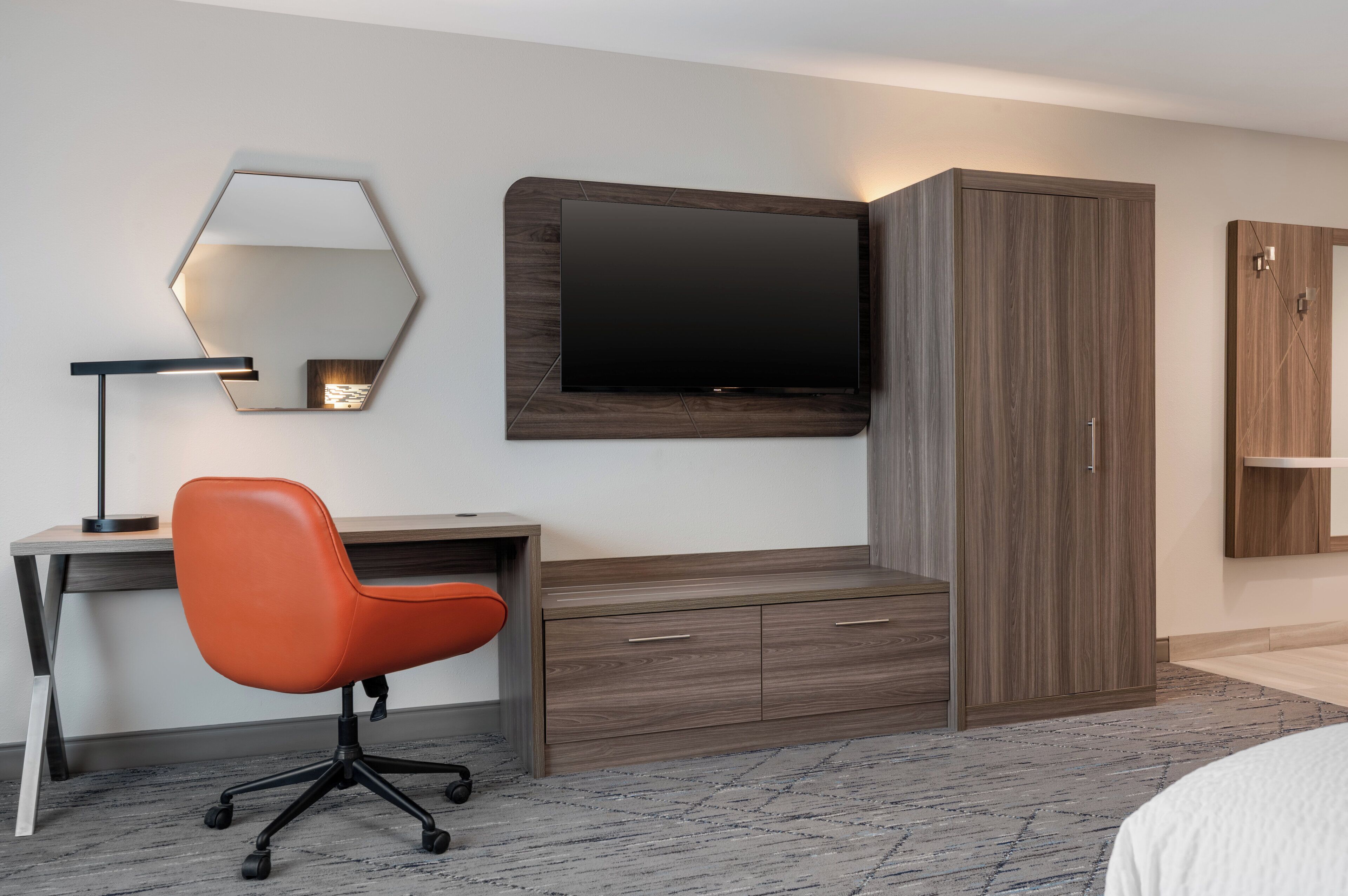 Una habitación de hotel moderna con un escritorio de madera, una silla de oficina naranja, un televisor de pantalla plana grande montado en la pared y un armario con espejo.