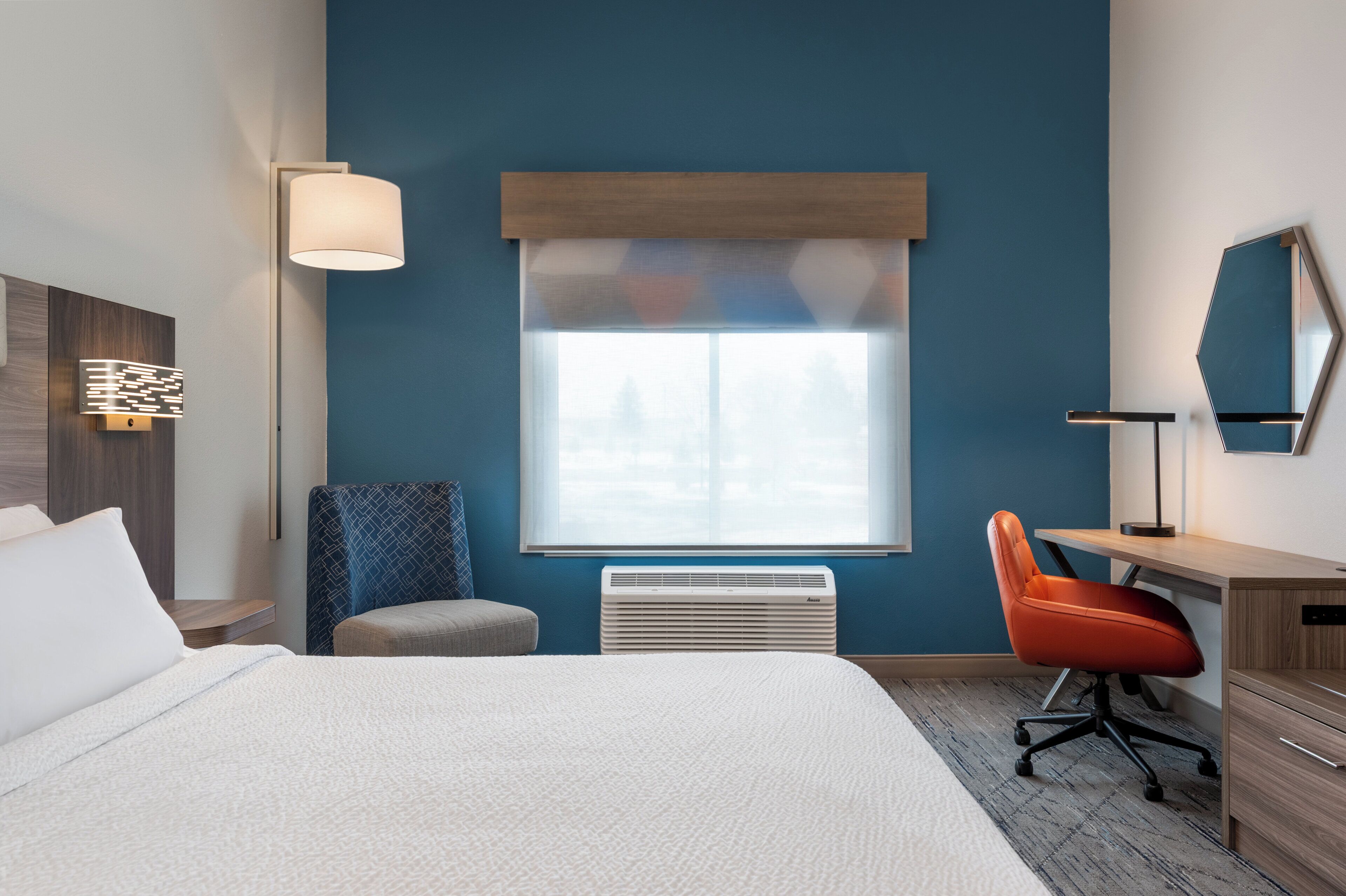 A hotel room with a large bed, a desk with a chair, a window with blinds, a wall-mounted lamp, and a heater.