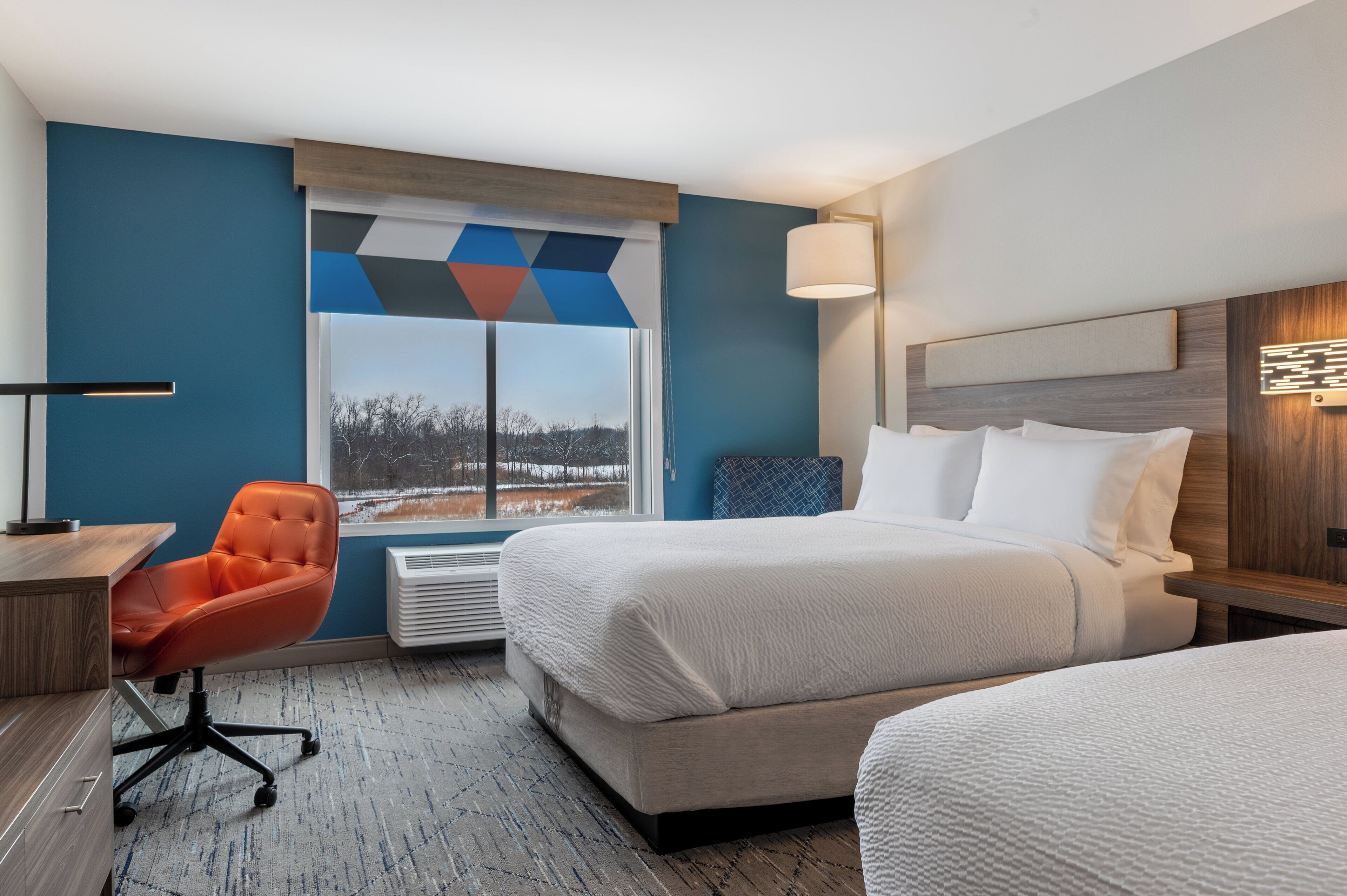 Habitación de hotel con una cama grande, un escritorio con silla, una ventana con vistas a árboles y una lámpara fijada a la pared.