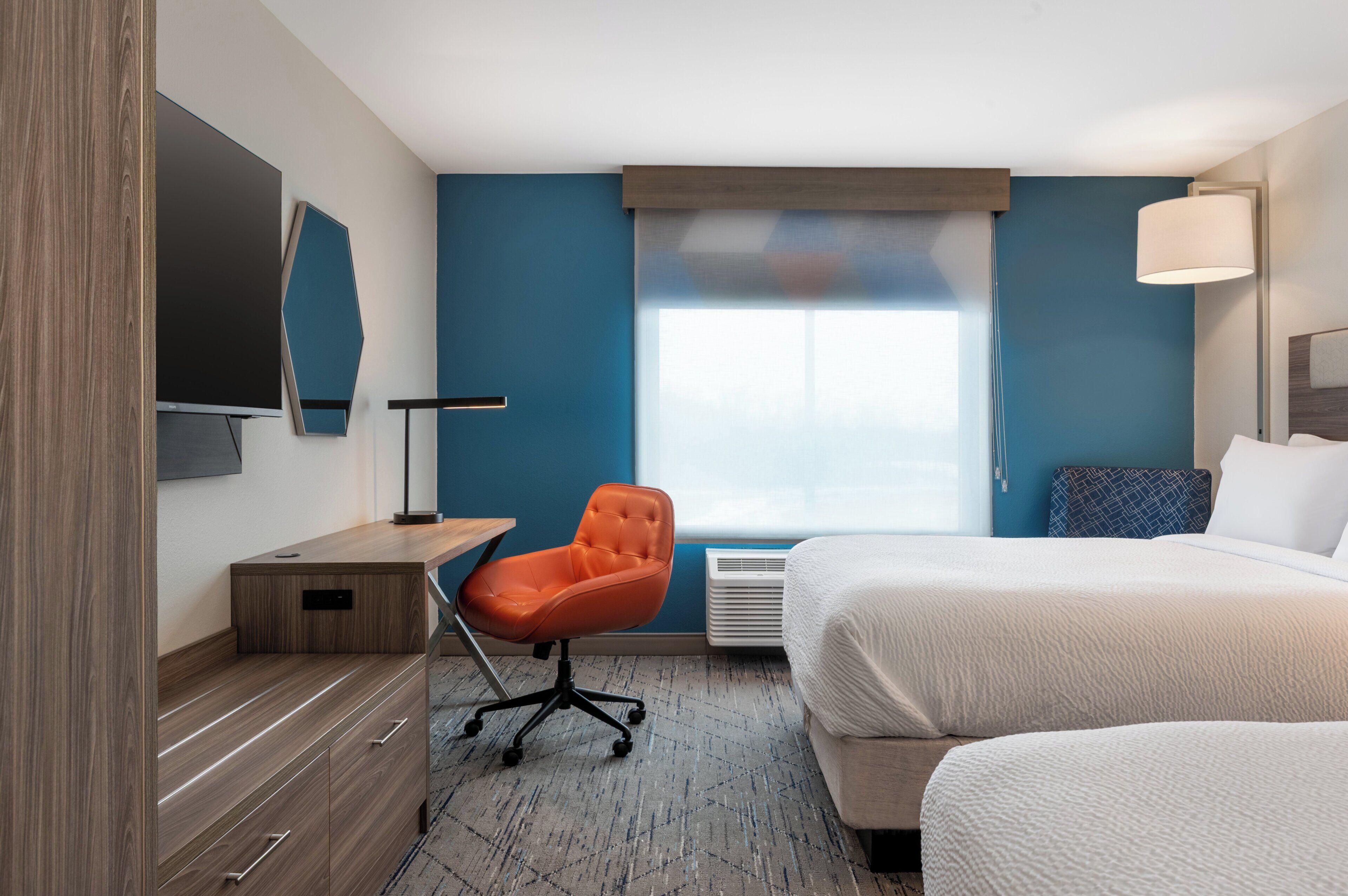 Una camera d'albergo con un letto, una scrivania con sedia, un televisore a schermo piatto e un'ampia finestra con tende.