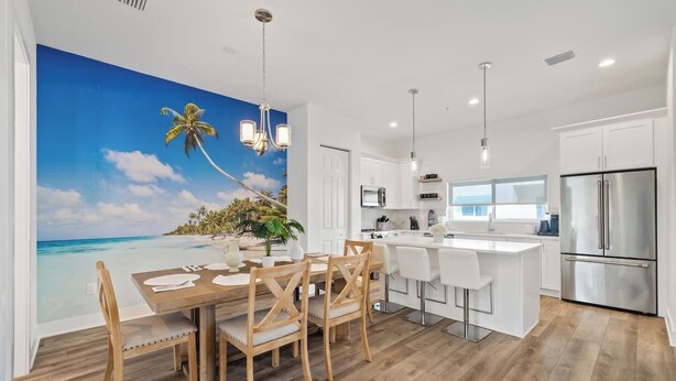 Una cocina moderna con armarios blancos, un refrigerador de acero inoxidable y una zona de comedor con una mesa y sillas de madera.