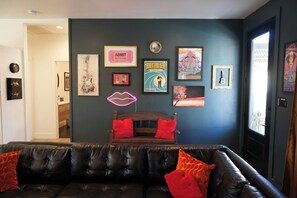 Een kamer met een donkerblauwe wand, een zwarte leren bank met rode kussens, en diverse lijstwerken en posters.