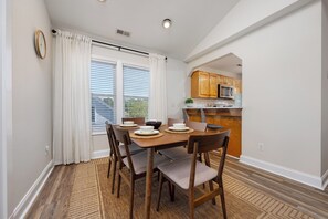 Un comedor con una mesa y sillas de madera, una cocina al fondo y una ventana con persianas.