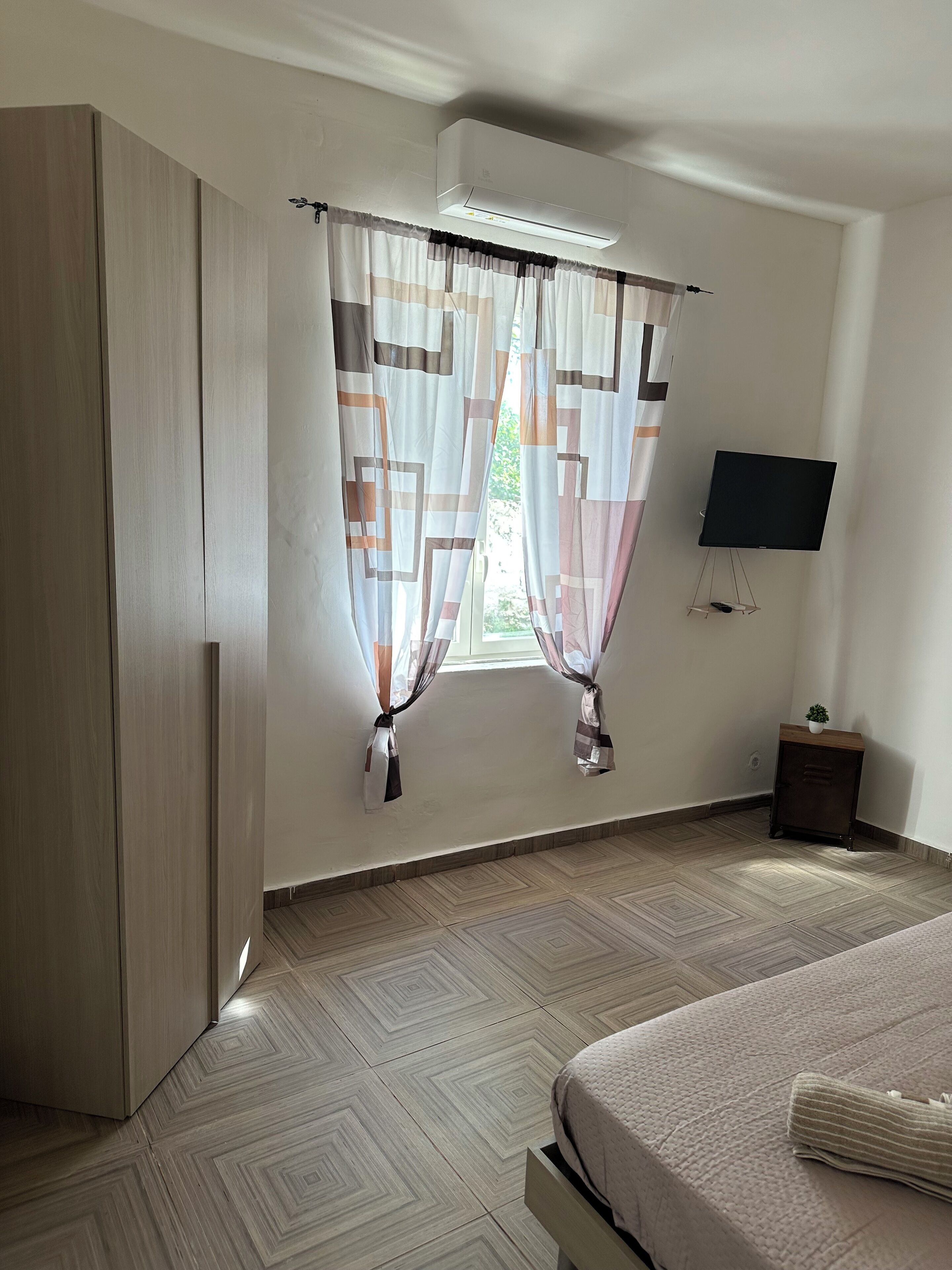 A bedroom with a bed, a wardrobe, a TV on a stand, and a window with curtains.