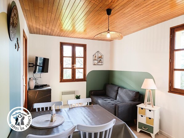 Une pièce chaleureuse avec un plafond en bois, un canapé vert, une table à manger avec des chaises, un téléviseur et une fenêtre donnant sur un beau paysage.