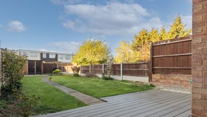 A backyard with a wooden deck, a grassy lawn, and a brick wall. - Spacious 3-Bedroom Home in Basildon, Essex London (Basildon)