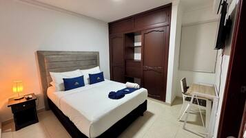 A neatly made bed with white and blue bedding, a wooden headboard, and a bedside table with a lamp. There is a wooden wardrobe and a small desk with a chair in the room.