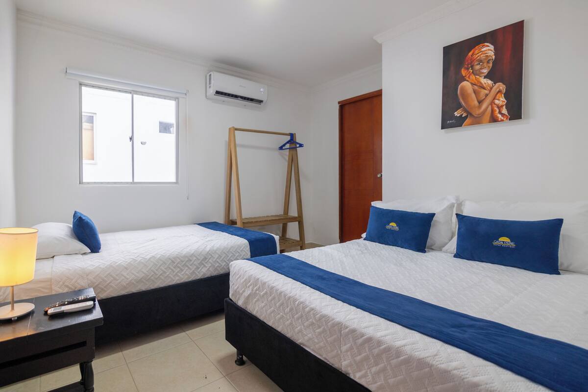 Quarto de hotel com duas camas, um cabide de madeira, um quadro na parede e uma janela com persianas.