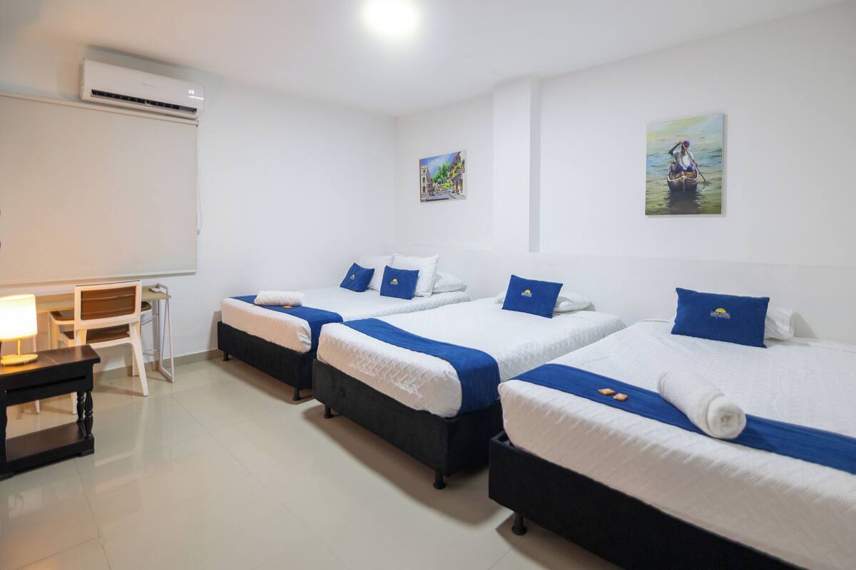Quarto de hotel com três camas, cada uma com roupa de cama branca e azul, uma escrivaninha com abajur e ar condicionado na parede.