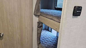 A compact RV interior with a wooden door, a bunk bed with a patterned mattress, and a small cabinet.