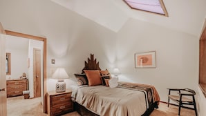 A bedroom with a skylight, a bed with a patterned bedspread, a nightstand with a lamp, a wooden dresser, and a chair. - Condo 91sw With a River View and Close to Community Amenities (Deming)