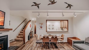A modern living room with a fireplace, wooden flooring, a dining area with a table and chairs, and a staircase with a wooden railing. - Condo 91sw With a River View and Close to Community Amenities (Deming)