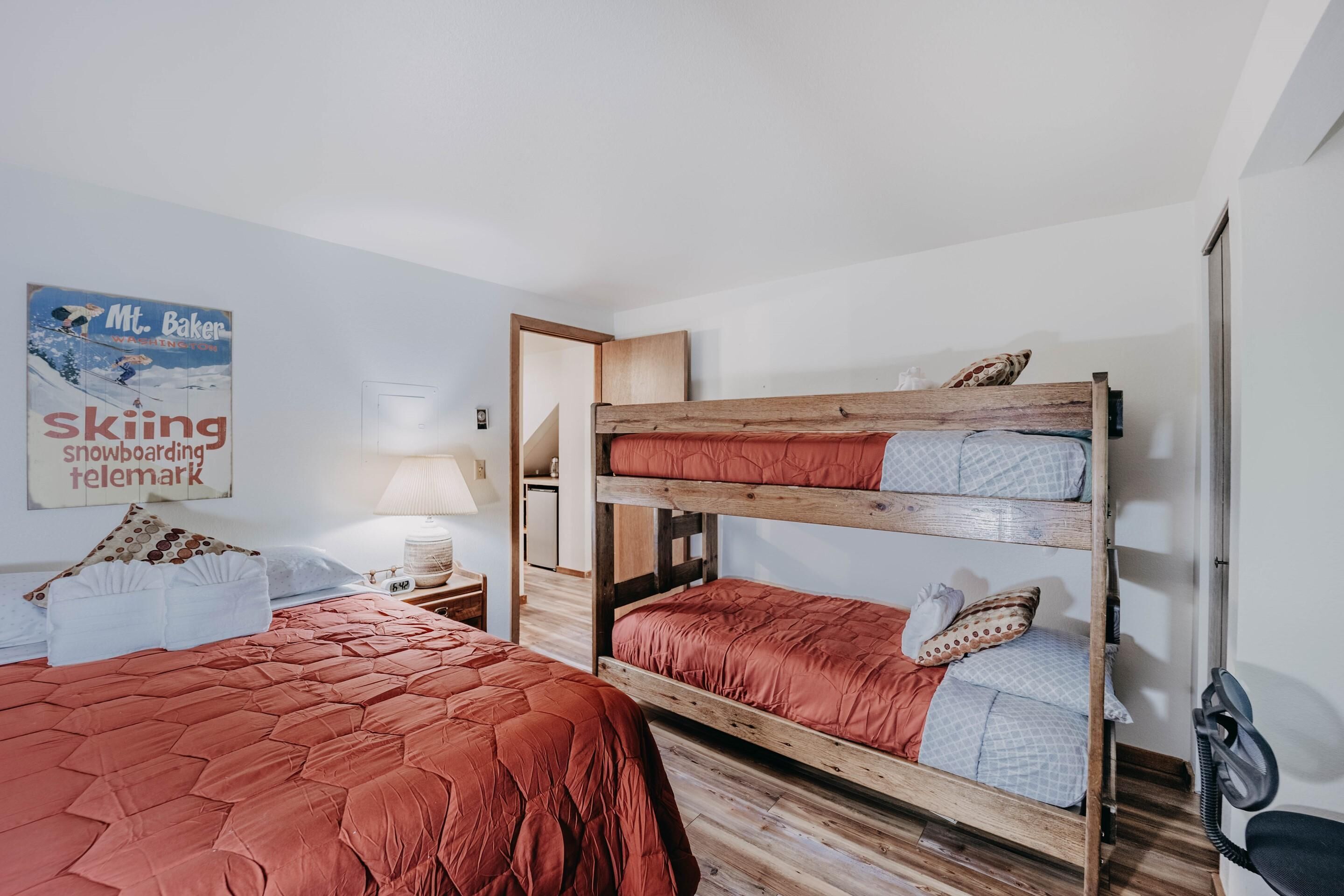 Habitación con dos camas, una con colcha roja y la otra con colcha azul. Se observa una cama nido de madera con colcha roja y azul, un cartel en la pared y una lámpara en la mesita de noche.