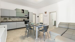 A modern kitchen with grey cabinets, a microwave, and a tiled backsplash. There is a dining table with chairs and a sofa in the living area. - Alguerìta Apartment - Alghero (Alghero)