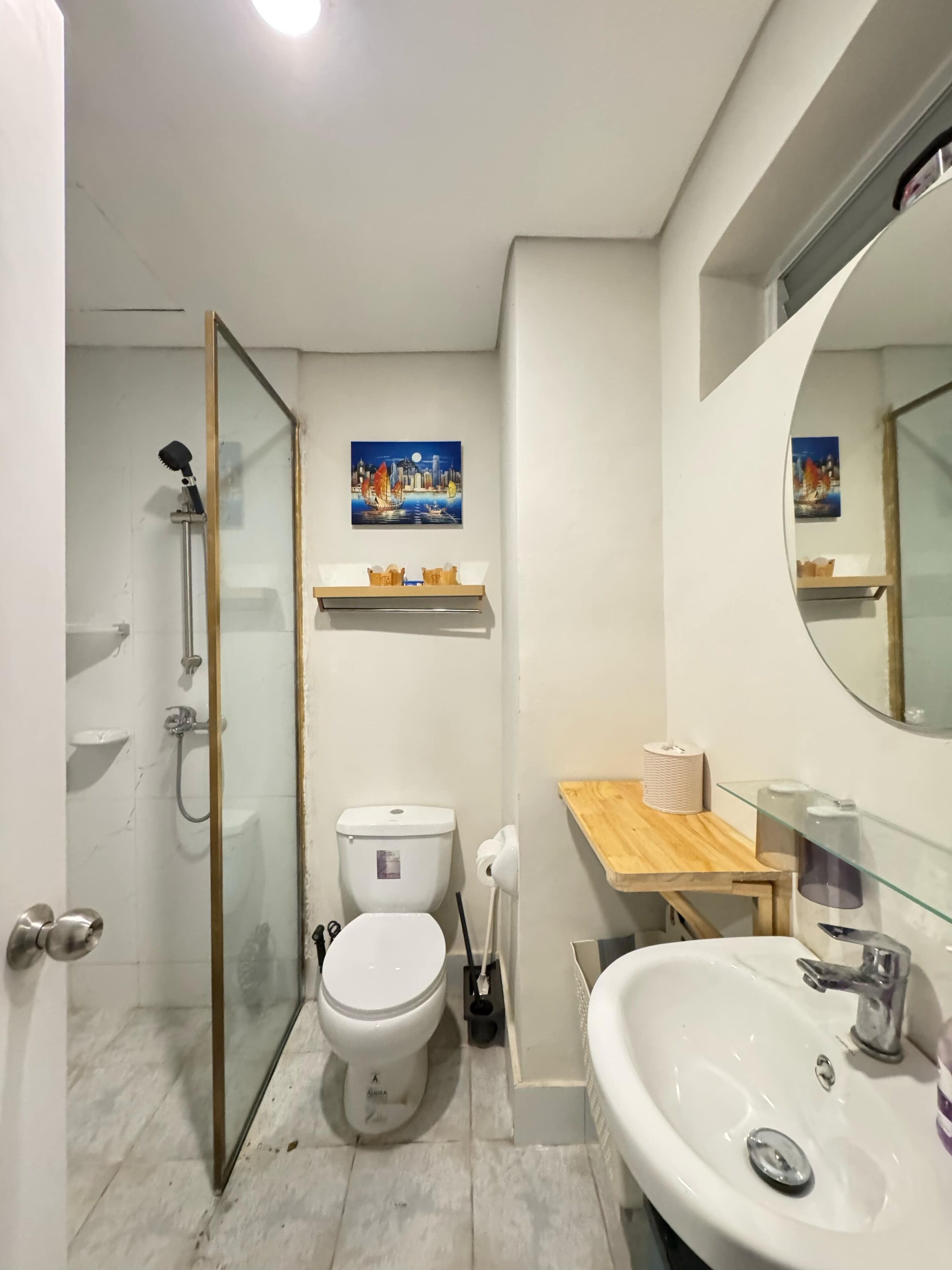 A bathroom with a glass shower enclosure, a toilet, a sink, and a mirror. There are two wall-mounted shelves with toiletries and a painting above the toilet.