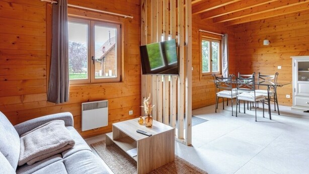 A wooden cabin interior with a sofa, a TV mounted on a wall, a dining table with chairs, and a window with curtains. - "Domaine la Clairière Le Douglas (Soultzmatt)