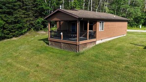 Eine kleine, einstöckige Holzhütte mit Veranda und einem umzäunten Deck, umgeben von einer Wiese und dichtem Wald.