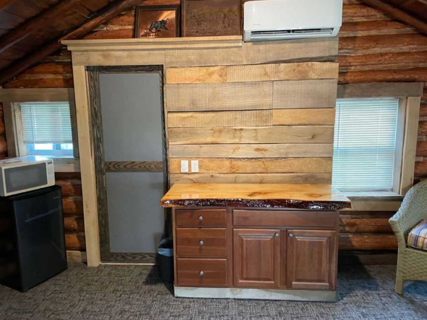 Interior rústico de una cabaña con una pared de madera, una pequeña kitchenette con microondas y una ventana con persianas.