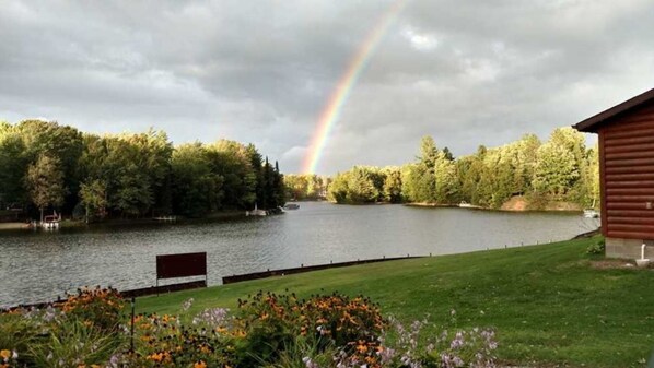 Vista de un lago con un arcoíris, un muelle de madera y un área verde con flores.