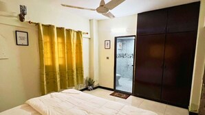 Une chambre à coucher avec un lit, un ventilateur de plafond, une armoire et une salle de bain visible à travers une porte ouverte.