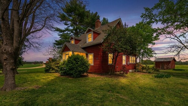 Une maison en bois rouge, avec un grand arbre au premier plan, entourée d’une pelouse bien entretenue et d’un petit abri de jardin à l’arrière-plan.