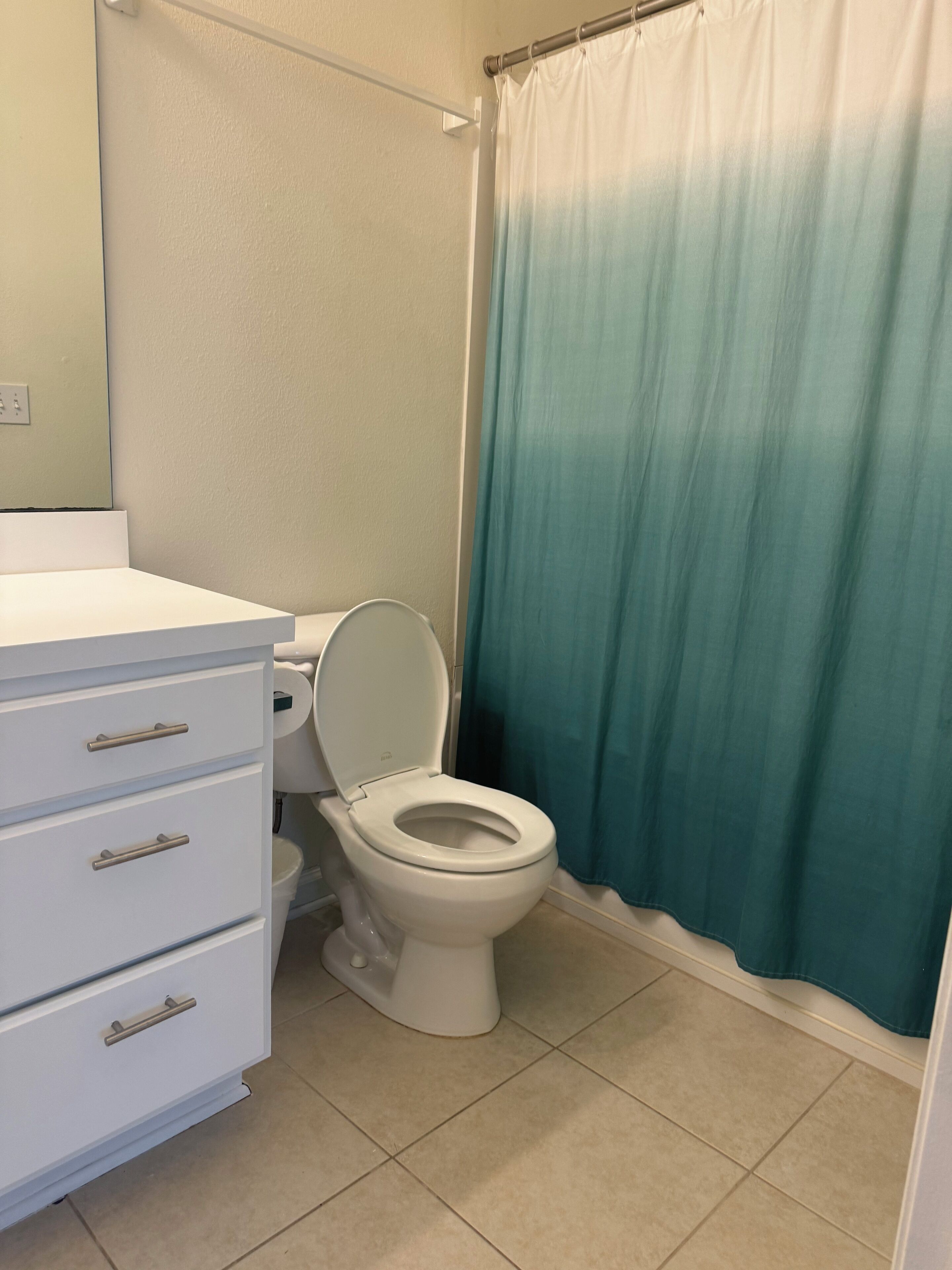 Un baño con un inodoro blanco, un armario blanco con cajones y una cortina de ducha color turquesa.